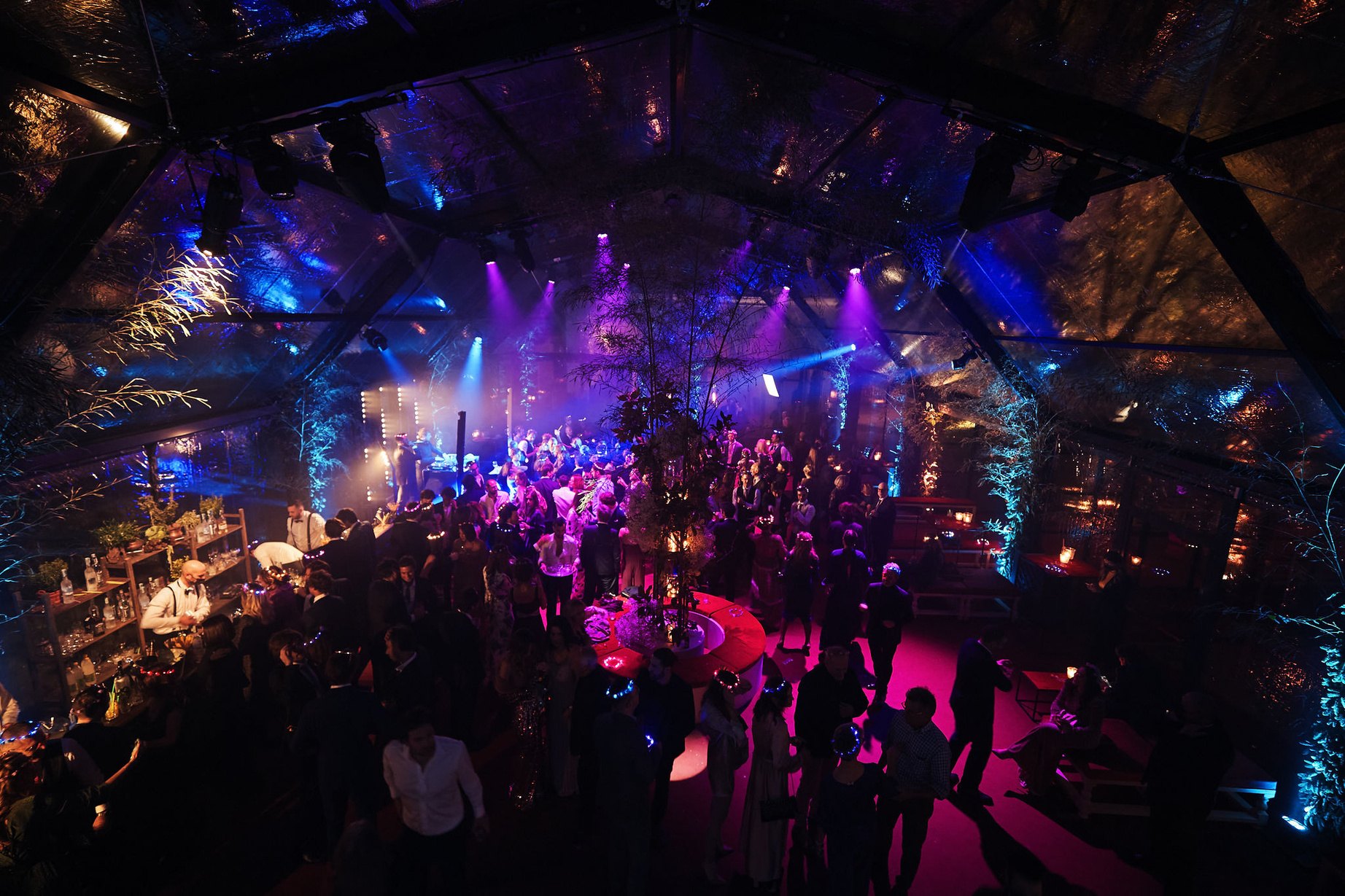 Veduta dall'alto di una festa serale dentro una struttura con copertura trasparente, la sala piena di ospiti e luci colorate